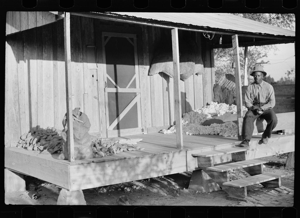 ชาวไร่ผิวดำนั่งบนระเบียงไม้หน้าบ้านพัก ชานหน้าบ้านมีฝ้ายกับหัวมันเทศกองอยู่ ภาพนี้ถูกถ่ายในปี ค,ศ, 1939 (Library of Congress, LC-USF33- 030572-M2)
