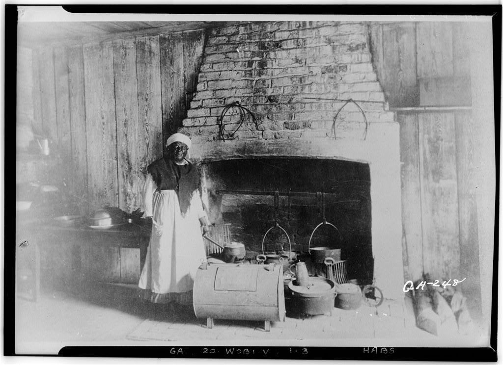 แม่ครัวผิวดำยืนหน้าเตาไฟในห้องครัว ช่วงปลายคริสตศตวรรษที่ 19 (Library of Congress, HABS GA,20-WOBI.V,1--3)